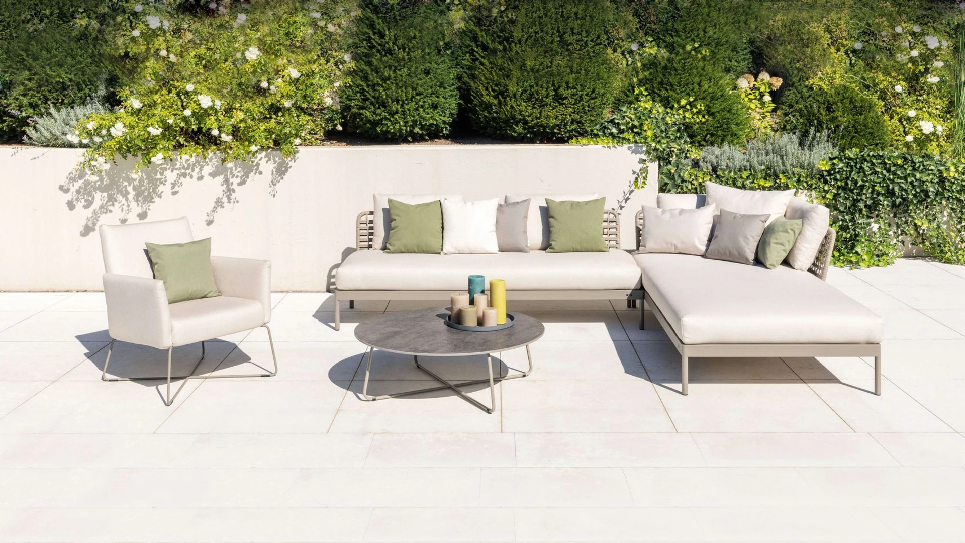 Helle Kairos Ecklounge und Kalos Sessel mit olivgrünen Akzentkissen. Moderne Outdoor-Möbelgruppe mit filigranen Gestellen vor einer begrünten Gartenmauer.