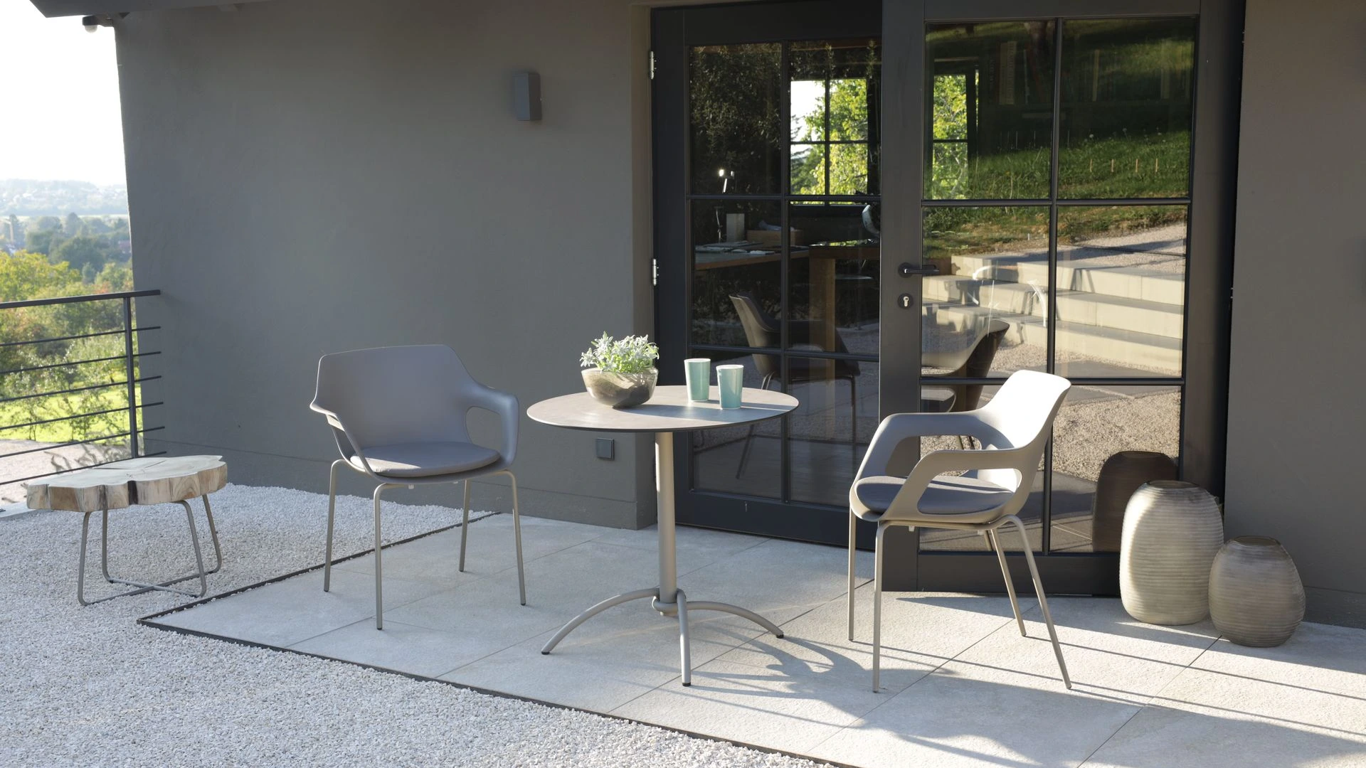 Tempus Schalensessel in Taupe mit Greystone-Gestell kombiniert mit einem Taku Esstisch; im Hintergrund ein Drop Tisch mit Unikat Teakholzbaumkante.