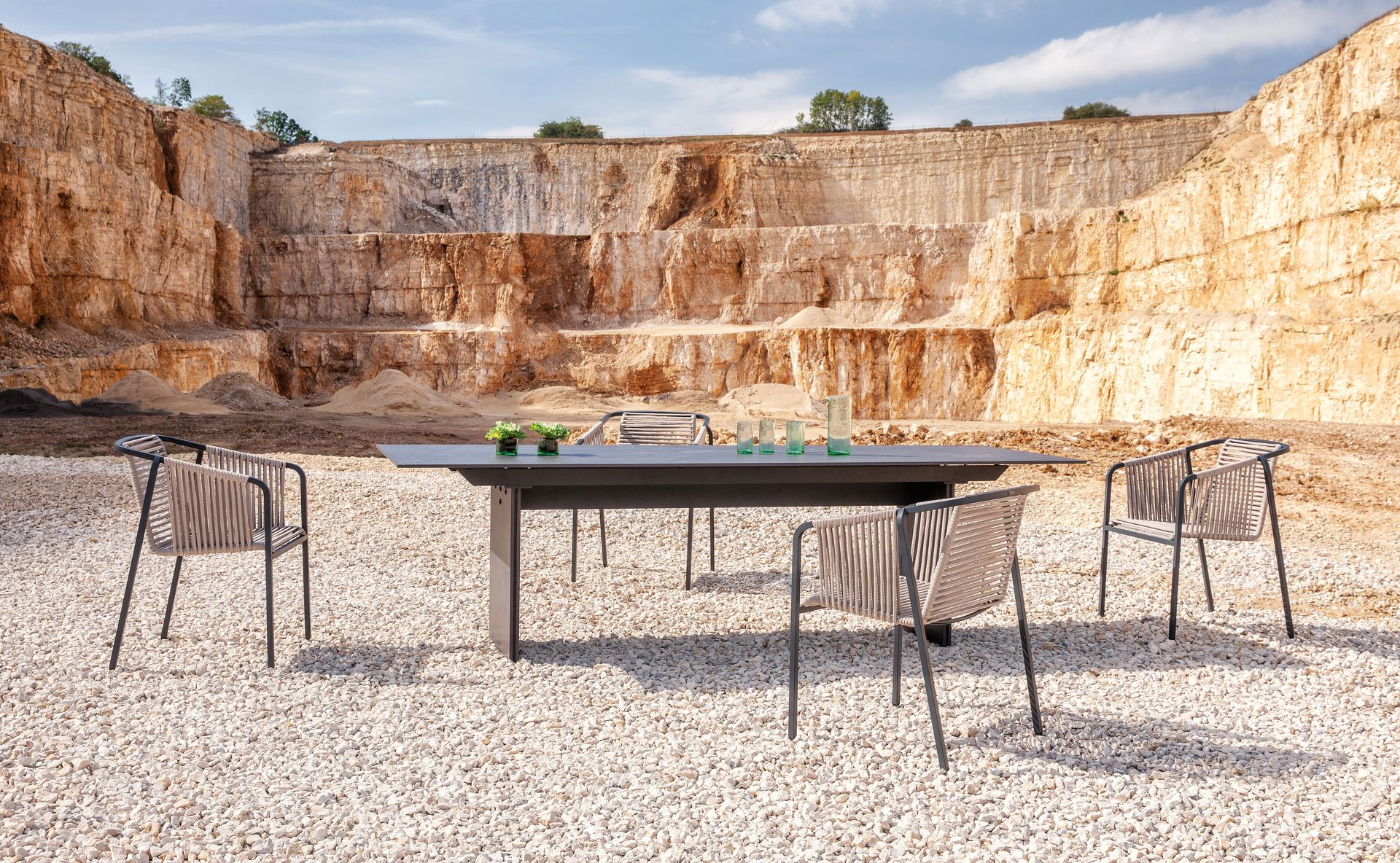 Ein Suite Sessel mit dunklem Gestell, dunkler Seilbespannung und hellgrauen Kissen neben einem Doppio Tisch bei einem Fotoshooting in einem Steinbruch.